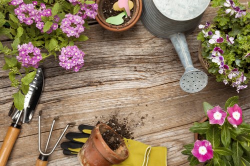 The image depicts a close-up view of a garden workspace on a wooden surface, with various gardening tools and potted plants. In the top left corner, there are clusters of pink and purple flowering plants in small pots, adding vibrant colour to the scene. To the right, a grey watering can with a perforated spout is positioned alongside a small clay pot filled with garden chalk and plant markers. On the left side, garden tools including a hand rake, trowel, and pruning shears are laid out, with soil scattered nearby. A pair of yellow gardening gloves lies in front of a small empty terracotta pot, ready for planting. The wooden surface and surrounding potted plants suggest an outdoor gardening environment, possibly a backyard or patio area, with natural daylight illuminating the scene. This setup reflects typical preparations for garden planting, potting, and garden bed maintenance, aligning with gardening and landscaping services offered by Landscapers Enfield in the local Enfield area.