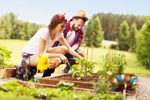 A woman and a man are working together in a garden bed situated in a spacious outdoor area, with the woman planting or tending to small, leafy green plants while the man watches and assists nearby. The garden bed is bordered by wooden planks, and there are various garden decorations, including a yellow chick ornament and a small clay pot painted with bright colors, placed on the soil. In the background, lush green trees and a well-maintained lawn extend into the distance under a clear, sunny sky. The scene suggests a peaceful, well-kept garden environment suitable for outdoor planting and lawn care, reflecting typical features of a landscaped backyard or front garden in Enfield. The natural textures of the soil, the vibrant green of the plants, and the cheerful outdoor atmosphere highlight the gardening activity, which a professional landscaper from Landscapers Enfield might support through planting and landscaping services.