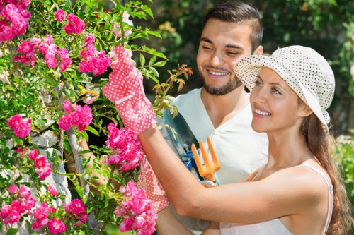 A smiling young couple working together in a lush, well-maintained garden, with the woman wearing a white polka-dot hat and gardening gloves, tending to vibrant pink flowering bushes, while the man, dressed casually, assists nearby. The garden features a mix of green foliage, flowering plants, and a neatly trimmed lawn, with sunlight filtering through the trees in the background. The scene reflects outdoor maintenance and gardening activities typical of Enfield, with a focus on flower care and garden upkeep, supported by professional landscaping services such as those from Landscapers Enfield, highlighting a tidy, flourishing outdoor space.