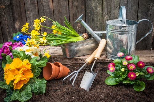 A selection of garden tools and flowers arranged on dark soil in a backyard setting. Visible elements include a metal watering can, a small gardening fork, a trowel, and a hand rake, all positioned on the freshly turned soil. To the left, there are bright yellow and purple primroses in full bloom, with some pink and white flowers nearby. Behind the flowers, a small terracotta plant pot lies on its side. The background features a weathered wooden fence, indicating this is a private garden space, likely in Enfield. The scene suggests preparations for planting or ongoing garden maintenance during daylight hours with overcast weather, creating a natural, outdoor environment suitable for gardening services by Landscapers Enfield. The overall composition highlights the vibrant colors of the flowers, the varied textures of garden tools, and the neat landscaping layout, reflecting professional outdoor care and gardening work typical for the area.