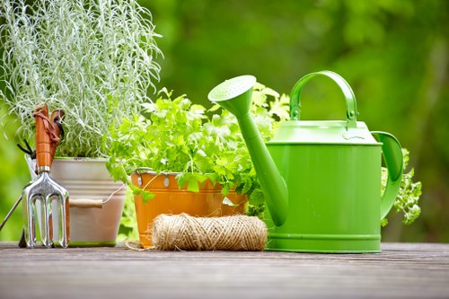 A close-up view of a well-maintained garden outdoor space, featuring a weathered wooden surface in the foreground with various gardening tools and supplies arranged neatly. On the left, there are two potted plants: a tall, silver-grey herb with fine, feathery foliage and a smaller terracotta pot containing vibrant green leafy herbs. In the center, a coiled natural hemp garden twine is positioned beside a bright green plastic watering can with a long spout. The background reveals lush, blurred greenery with out-of-focus trees or shrubs, suggesting a flourishing garden environment typical of outdoor maintenance in Enfield or the surrounding areas. The overall scene depicts a tidy and organized gardening setup, highlighting essential tools and healthy plants, suitable for landscaping or garden care services offered by Landscapers Enfield.