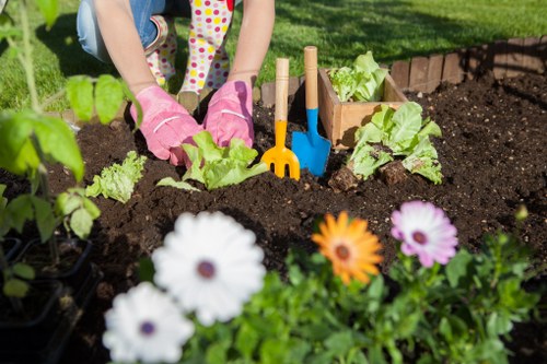 The image depicts a person planting young lettuce plants into a prepared garden bed with rich, dark soil in an outdoor setting. The gardener's hands are visible, wearing pink gardening gloves, and they are using small blue and orange garden tools to assist with planting. In the foreground, there is a cluster of colourful flowering plants, including white, purple, and orange daisy-like blossoms, adding vibrant colour contrast to the garden scene. The background showcases a well-maintained lawn with lush green grass and a neatly edged flower bed bordered by bricks, creating a tidy garden layout typical of local residential outdoor spaces. Natural sunlight illuminates the scene, indicating a clear, bright day suitable for gardening activities. This scene captures the essential elements of gardening and planting work, reflected in the professional landscaping services offered by companies such as Landscapers Enfield, who focus on outdoor beautification and maintenance within the Enfield area.