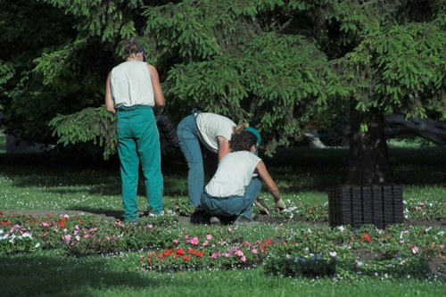Two gardeners are working in a lush outdoor garden space, surrounded by dense greenery and tall trees with vibrant green foliage. One gardener, standing and wearing teal trousers and a sleeveless top, observes while the other two, one kneeling and the other bent over, tend to a flower bed. The flower bed contains a variety of small, colorful flowering plants with pink, red, and white blossoms, arranged along the edge of a well-maintained, dense green lawn. A black grid-style garden edging border is visible on the right side of the image, separating the flower bed from the grass. The scene is illuminated by natural daylight, suggesting a bright, overcast or partly cloudy day, typical of outdoor gardening activities in Enfield. The setting appears to be a garden or backyard, with natural textures of soil, grass, and foliage clearly visible, highlighting professional gardening care by Landscapers Enfield in an English suburban environment.
