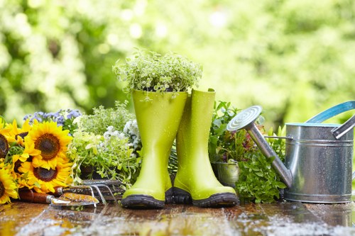 A close-up view of a gardening workspace outdoors, featuring a pair of bright green rubber rain boots used as a flower container filled with white blossoms on a wooden table. Surrounding the boots are potted plants with small green foliage and colorful flowers, including yellow sunflowers and purple blooms. To the right, there is a silver metal watering can with a long spout and a blue handle, and gardening tools are also visible, suggesting active outdoor gardening. The background shows blurred lush green trees and foliage, indicating a well-maintained private garden or backyard space in Enfield, with natural daylight and a fresh, vibrant atmosphere conducive to plant care and landscaping work.