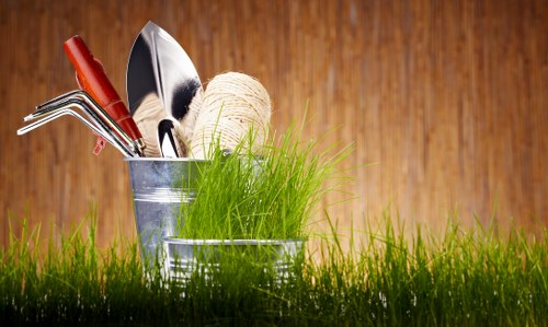 A close-up image of a garden scene in a residential outdoor space, featuring a patch of well-maintained, vibrant green grass in the foreground. Behind the grass, two small metal containers are visible, one holding gardening tools including a small hand rake and trowel with red handles, and the other containing a ball of twine and a tuft of fresh, bright green grass or young plants. The background showcases a warm-toned wooden fence or wall, suggesting a tidy and organized garden environment typical of landscaped yards in Enfield. The overall scene emphasizes gardening tools and lively plant elements suitable for outdoor maintenance and lawn care services offered by landscapersenfield.co.uk, aligned with their focus on sustainable and eco-friendly gardening practices. The lighting indicates bright natural daylight, contributing to a fresh and inviting appearance consistent with professional garden care.