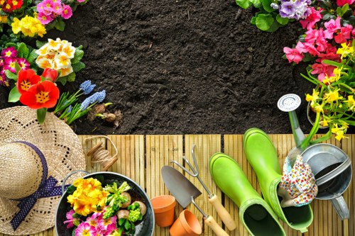 The image shows a well-maintained garden area with a wooden decking surface in the foreground, surrounded by various colorful flowering plants such as primroses, pansies, and other seasonal blooms along the edges. To the right, there are bright green rubber boots placed beside a small gardening trowel, a hand fork, and a watering can filled with soil and plants, indicating active gardening work. A straw sun hat and a decorative flower pot are also visible on the decking, suggesting outdoor maintenance activities. Behind the garden elements, dark rich soil is prepared for planting, with the garden space enclosed by lush greenery and shrubbery. The scene is set in natural daylight, with a clear, slightly overcast sky, creating a soft light that highlights the vivid colors of the flowers and garden tools. This outdoor environment exemplifies typical garden setup and maintenance, tailored for a residential front or back yard in Enfield, aligning with professional gardening and landscaping services offered by Landscapers Enfield.