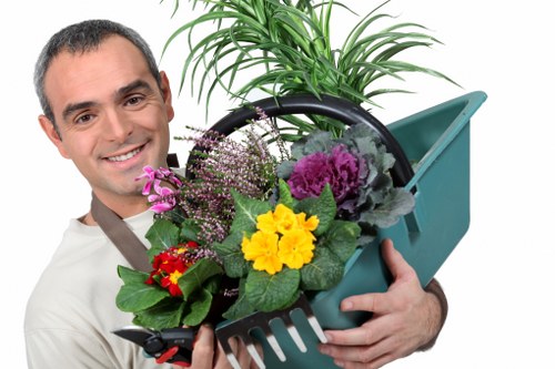 A smiling man with short dark hair, dressed in a light-colored shirt, holding a green plastic gardening trowel and a blue watering can in an outdoor garden setting. The garden features a well-maintained lawn in the foreground with lush, green grass. Behind him, there are small shrubs and plants, with a large, leafy potted plant placed near a paved or decking area. The weather appears to be bright and clear, suggesting a sunny day, suitable for gardening activities. This image reflects professional gardening and landscaping practice, suitable for a gardening service website such as landscapersenfield.co.uk, emphasizing outdoor maintenance and garden care in the Enfield area.