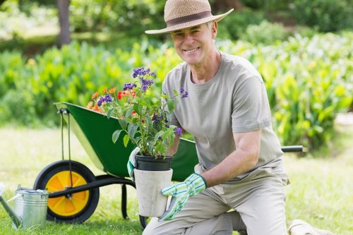 The image shows an elderly man kneeling on a well-maintained lawn in a garden, holding a potted plant with purple and red flowers, while smiling at the camera. Behind him, there is a green garden wheelbarrow with a bright yellow wheel, filled with gardening supplies, and a metal watering can resting on the grass nearby. The garden features a lush, green hedge in the background, along with various shrubs and trees, indicating a private outdoor space in a suburban setting. The weather appears sunny with clear lighting, suitable for outdoor gardening tasks. The man is dressed in light-colored gardening clothes, including a wide-brimmed hat for sun protection and green-striped gardening gloves. The scene conveys a peaceful and neatly landscaped garden environment, highlighting elements typical of professional gardening and landscaping services, such as lawn care, planting, and outdoor maintenance, relevant to Enfield or nearby areas in North London.