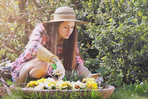 A young woman tending to a flower bed in a well-maintained garden, with lush green foliage and dense bushes in the background. She is wearing a straw hat, a plaid shirt, and gardening gloves, and is carefully planting or tending to a variety of colorful flowers in the soil within a circular flower bed edging. The garden features a neatly cut grassy lawn, and the scene is illuminated by natural sunlight, indicating a bright and clear day. This outdoor space exemplifies typical residential garden landscaping, suitable for regular maintenance and gardening services by companies like Landscapers Enfield in the Enfield area, with a focus on creating vibrant and managed garden environments.