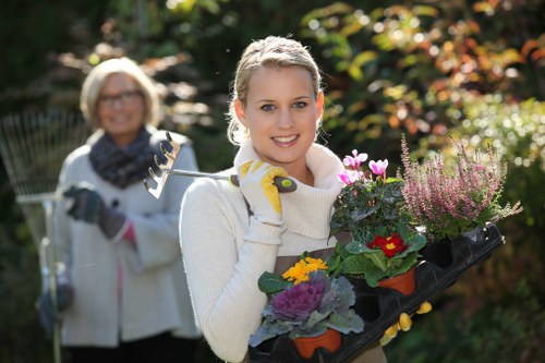 A young woman with blonde hair, wearing a white sweater and gardening gloves, is standing outdoors in a garden during daylight, holding a black tray filled with various colorful flowering plants and foliage, including red, yellow, and purple blossoms with green leaves, in front of a lush garden background with dense shrubbery and trees. In the blurred background, an older woman with glasses and a scarf is also gardening, holding a pruning tool, amidst the greenery. The scene is well-lit by natural sunlight, highlighting the vibrant colors of the plants and contributing to a bright, garden-friendly atmosphere that reflects professional gardening and landscaping activities typical of a landscaped outdoor space in Enfield, UK.